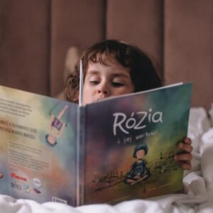 Dziecko siedzi na łóżku i czyta książkę „Rózia i jej wartości.